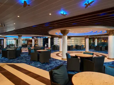 Elegante Bar-Lounge auf der Carnival Venezia. Blauer Teppich, Korbsessel, runde Tische, Säulen, blaue Deckenbeleuchtung. Bar und Bühne im Hintergrund. Ohne Personen.