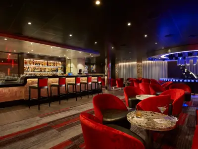 Blick in eine schicke Bar-Lounge auf der Carnival Venezia mit einer langen Bar, roten Samtsesseln und kleinen Tischen, im Hintergrund eine Bühne mit einem stilisierten Klavier unter blauer Beleuchtung.