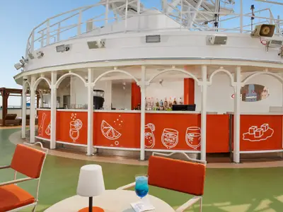 Außenbar auf der Carnival Venezia. Orange Theke mit Drink-Grafiken, weiße Sessel mit orangen Polstern, Tisch mit blauem Drink. Blauer Himmel.