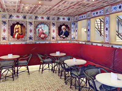 Eine Innenansicht einer Lounge auf der Carnival Venezia mit einer durchgehenden, roten Sitzbank, runden Tischen und Korbstühlen. Die Wände sind mit gerahmten, humorvoll-modernisierten Porträts klassischer Persönlichkeiten verziert. Ein Deckenfenster zeigt eine Treppe.