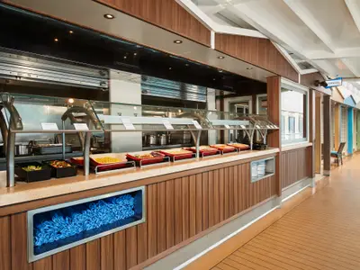 Ein modernes Buffet auf der Carnival Venezia mit einer langen Theke voller warmer Speisen. Der Bereich ist mit Holzpaneelen und einem hellen Arbeitsbereich gestaltet. Blaue Servietten sind in einem Fach unten sichtbar und im Hintergrund sind Sitzgelegenheiten an einer Promenade zu erkennen.