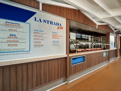Detailansicht des Buffetbereichs 'LA STRADA grill' auf der Carnival Venezia. Eine große Speisekarte ist links zu sehen, daneben eine Selbstbedienungstheke mit verschiedenen Gerichten in Wannen. Die Theke hat eine Holzverkleidung und blaue Lichtinstallationen.