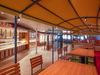 Weitwinkelaufnahme eines modernen Buffetrestaurants auf der Carnival Venezia mit einer langen Theke, Essensauslagen, leeren Tischen und bequemen Sitzbänken unter einem markanten, orangefarbenen Stoffdach. Eine runde Metallstruktur steht im Zentrum des Raumes.