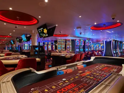 Ein geräumiges Casino auf der Carnival Venezia, beleuchtet von roten und blauen Lichtern. Zahlreiche Slot-Maschinen und Spieltische wie Craps und Kartentische mit roten Stühlen sind zu sehen.