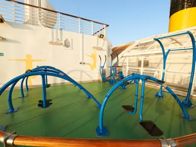 Outdoor-Fitnessbereich auf der Carnival Venezia mit verschiedenen blauen Trainingsgeräten auf grünem Boden. Die weißen Wände zeigen gelbe Silhouetten von sportlichen Personen.