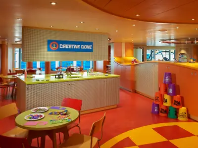 Bunter Kinderbereich an Bord der Carnival Venezia. Im Vordergrund ein grüner Tisch mit Bastelmaterial. Ein großes Schild mit "Creative Cove" ist sichtbar. Im Hintergrund Fenster und weitere Tische. Eine Regalwand mit Spielzeug, darunter ein Turm aus bunten Bechern, rundet das Bild ab.