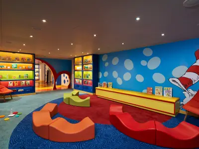 Farbenfroher Kinderbereich auf einem Schiff. Blaue Wände mit Dr. Seuss Motiv, bunte Bücherregale, weiche Sitzmöbel und Spielzeug auf Teppichboden.