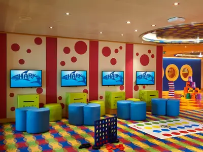 Ein farbenfroher Innenraum für Kinder auf der Carnival Venezia. Mehrere Fernseher zeigen das "SHARK"-Logo. Blaue und grüne Sitzpuffs, sowie Bodenspiele wie Twister und Vier gewinnt, laden zum Spielen ein. Im Hintergrund sind runde Fenster zu sehen.