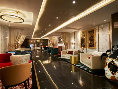 Eine luxuriöse Lounge an Bord der Carnival Venezia, geschmückt mit Marmorwänden, goldenen Akzenten und einer Vielzahl von farbigen Sitzbereichen. Ein großer Kronleuchter hängt von der Decke, und zwei Kunstwerke mit goldenen Masken zieren die rechte Wand.