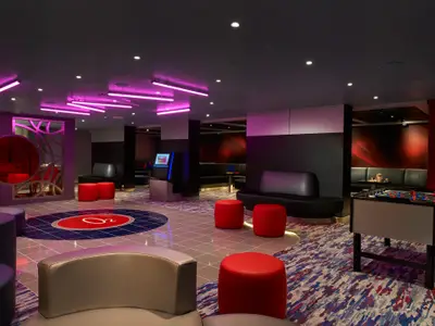 Ein moderner, dunkel gehaltener Loungebereich auf der Carnival Venezia mit roten Sitzhockern, schwarzen Sofas und einem Kicker. Lila Neonlichter an der Decke und ein großes O2-Logo auf dem Fußboden in der Mitte des Raumes.