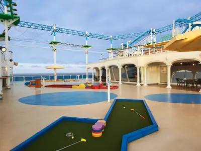 Ein weitläufiges Oberdeck der Carnival Venezia bei Tageslicht. Links eine Minigolfanlage, im Hintergrund ein Hochseilgarten. Das blaue Meer und der Himmel sind am Horizont sichtbar. Der Deckboden ist hellbeige mit blauen Kreisen. Mehrere weiße Strukturen, Pfeiler und Geländer sind zu sehen.