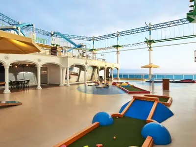 Ein heller Minigolfplatz und ein Hochseilgarten auf dem oberen Deck der Carnival Venezia. Im Hintergrund ist das blaue Meer unter klarem Himmel zu sehen. Elegante weiße Architektur mit Säulen praegt den überdachten Bereich.