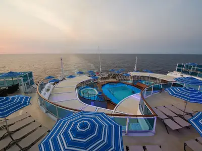 Das Heck der Carnival Venezia bei Sonnenuntergang. Großes Pooldeck mit mehreren Pools, Whirlpools, Liegen und blau-weißen Sonnenschirmen. Meer im Hintergrund.