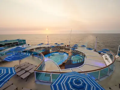 Weitwinkelansicht des Pooldecks der Carnival Venezia bei Sonnenuntergang. Ein ovaler Pool und ein Whirlpool sind von Liegestühlen und blau-weiß gestreiften Sonnenschirmen umgeben. Im Hintergrund ist das offene Meer mit tiefstehender Sonne sichtbar.