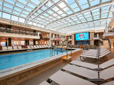 Innenansicht eines großzügigen Poolbereichs auf der Carnival Venezia mit großer Kinoleinwand am Kopfende, vielen Liegestühlen und einem verglasten Dach.