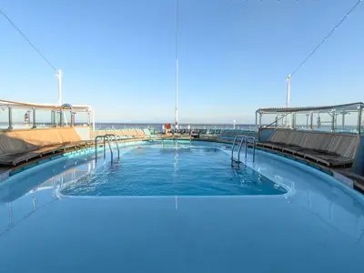 Breitwinkelaufnahme eines leeren Swimmingpools auf dem Oberdeck der Carnival Venezia bei Tageslicht. Das blaue Wasser des Pools reflektiert den wolkenlosen Himmel. Rund um den Pool befinden sich Holzliegen und Glasgeländer. Im Hintergrund ist das offene Meer zu sehen.