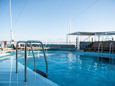 Detailansicht eines leeren, blauen Schwimmbeckens auf dem Sonnendeck der Carnival Venezia. Im Vordergrund sind polierte Edelstahlleitern zu sehen. Im Hintergrund liegen Sonnenliegen und Sitzbereiche unter einem klaren blauen Himmel.