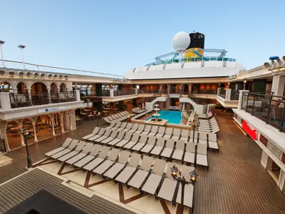 Blick von oben auf ein offenes Deck der Carnival Venezia. Ein rechteckiger Pool mit Brunnen, umgeben von Reihen beiger Liegestühle. Mehrere Ebenen mit Cafés und Balkonen sind sichtbar. Die Costa-Schornsteinspitze ist im Hintergrund erkennbar.