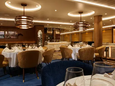 Elegantes Kreuzfahrt-Restaurant: Weiß gedeckte Tische, goldene Stühle und Gläser. Moderne Bar, Holzvertäfelung und Kunstwerke im Hintergrund.
