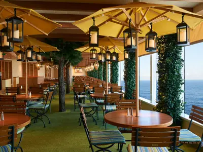 Geräumiges Schiffsrestaurant mit Holztischen und Stühlen, gelben Sonnenschirmen, Säulen mit grünen Pflanzen und Meerblick durch große Fenster. Beleuchtet von Laternen und Tageslicht.