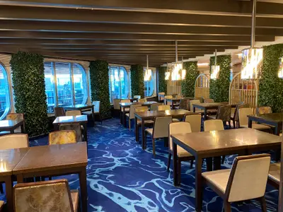 Ein leeres, modernes Restaurant auf der Carnival Venezia mit blauem Teppichboden, Holztischen und hellen Stühlen. Große Fenster mit Blick nach draußen sind von grünen Pflanzenwänden eingerahmt. Die Decke hat dunkle Holzbalken und moderne Hängelampen.