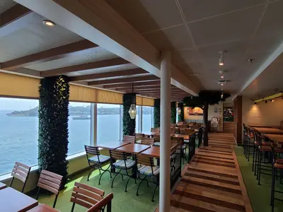 Geräumiges, helles Restaurant auf der Carnival Venezia mit großen Fenstern zum Meer. Grüne Teppichböden, Holztische und Stühle, sowie eine Reihe von Hochtischen und Bänken. Dekorationen umfassen Pflanzenwände und kunstvolle Hängelampen.