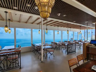 Helles Restaurant auf der Carnival Venezia mit raumhohen Fenstern und weitem Blick auf das blaue Meer und den Himmel. Elegante Pendelleuchten hängen von der Decke, während Reihen von Tischen und Stühlen den Raum füllen. Eine Servicetheke ist rechts sichtbar.