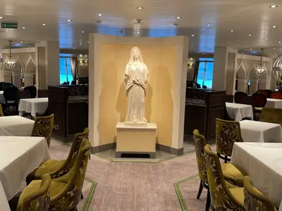 Ein elegantes Restaurant der Carnival Venezia mit festlich gedeckten Tischen, bequemen Stühlen und einer zentralen weißen Statue. Große Fenster bieten Blick nach draußen.