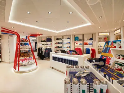 Ein heller, moderner Shop an Bord der Carnival Venezia, gefüllt mit Kleidung, Tassen und Souvenirs der Marke 'Carnival Venezia'. Eine markante rote und blaue Regalstruktur ist in der Mitte zu sehen. Die Regale sind sauber und übersichtlich präsentiert.
