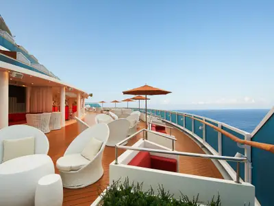 Weitwinkelaufnahme eines modernen Sonnendecks auf der Carnival Venezia mit weißen Rattan-Sesseln, Tischen, roten Sofas und orangefarbenen Sonnenschirmen. Im Hintergrund erstreckt sich das blaue Meer unter einem klaren Himmel.