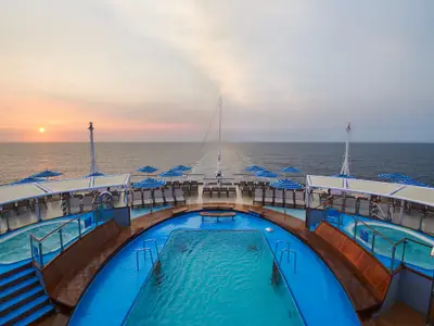 Sonnendeck der Carnival Venezia bei Sonnenuntergang. Ein großer Pool, zwei Whirlpools, Liegestühle und blau-weiße Sonnenschirme. Der Himmel über dem Meer ist warm orange gefärbt.