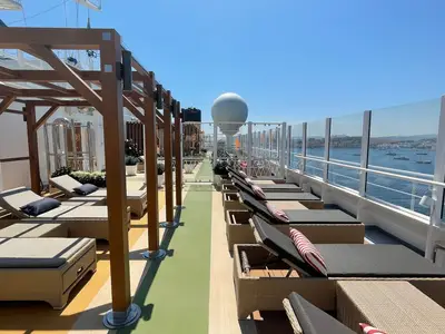 Blick über das Sonnendeck der Carnival Venezia an einem sonnigen Tag. Zahlreiche Liegestühle stehen unter einer Holzpergola und im Freien. Im Hintergrund ist das blaue Meer mit Yachten und eine Küstenlinie zu sehen.