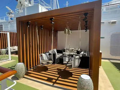 Eine moderne, überdachte Sitzecke auf dem Sonnendeck der Carnival Venezia. Zwei Schaukelstühle und eine Loungegruppe stehen auf Holz- und Kunstrasenboden, umgeben von weißen Relingen und einem weißen Schiffaufbau unter blauem Himmel.