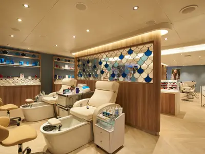 Heller, moderner Spa-Bereich auf der Carnival Venezia. Zwei Pediküre-Stationen, dekorative Trennwand mit blauen Muschelmotiven und ein Friseursalon. Warme Beleuchtung, Holzelemente.