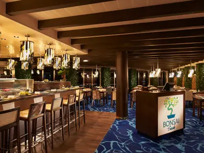 Innenansicht eines eleganten Bonsai Sushi Restaurants auf der Carnival Venezia. Links eine Sushi-Bar mit Auslage und Barhockern, rechts mehrere Tische mit Stühlen. Goldfarbene Design-Hängeleuchten und grüne Pflanzenwände schmücken den Raum. Der Teppich ist blau mit hellen Mustern.