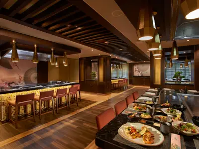 Asiatisches Spezialitätenrestaurant an Bord. Teppanyaki-Tische mit Speisen, Bar mit Hockern. Dunkles Holz, Gold-Akzente, Kirschblüten-Wandbild.