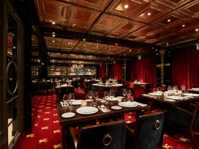 Ein luxuriöses Spezialitätenrestaurant an Bord der Carnival Venezia. Rote Samtvorhänge, ein goldener Deckenaufbau und dunkelbraune Tische mit weißem Geschirr und Gläsern schaffen eine festliche Atmosphäre. Im Hintergrund ist eine offene Küche mit Köchen zu sehen.