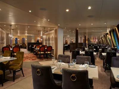 Ein elegantes, menschenleeres Spezialitätenrestaurant auf der Carnival Venezia. Dunkle Holzstühle und helle Tische sind festlich gedeckt. Die Decke ist mit Einbauspots beleuchtet und von Pendelleuchten in Kugelform akzentuiert. Die Wände zeigen bogenförmige Nischen und kunstvolle Wanddekorationen. Rechts sind große, schräg gestellte Fenster mit farbigen Vorhängen sichtbar. Der Boden ist mit gemusterten Teppichen und Parkett ausgelegt.