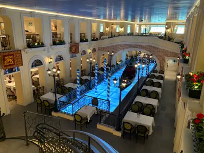 Blick von oben in ein thematisches Restaurant im Venedig-Stil mit Gondel, blau beleuchtetem Kanal und vielen Tischen mit Stühlen auf verschiedenen Ebenen.