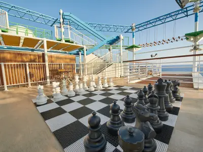Ein riesiges Schachspiel mit schwarzen und weißen Figuren auf einem schachbrettartigen Boden auf dem Oberdeck der Carnival Venezia. Im Hintergrund sind das Meer und ein Klettergarten zu sehen.