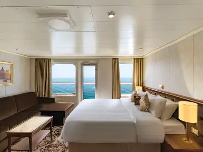 Elegante Suite der Carnival Venezia mit Doppelbett, braunem Sofa und Couchtisch. Große Fenster und eine Balkontür bieten weiten Blick auf das offene Meer.