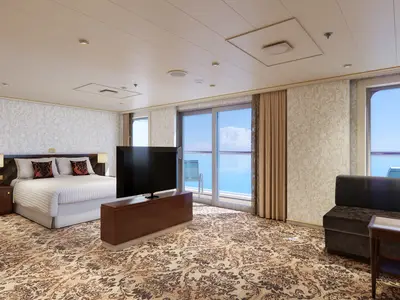 Geräumige Suite der Carnival Venezia mit großem Doppelbett, Nachttischen, einem Fernseher auf einem Sideboard, einem Sofa und einem Balkon mit Meerblick durch eine Glastür.
