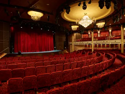 Blick in ein luxuriöses Theater auf der Carnival Venezia. Die Bühne ist mit einem roten Samtvorhang geschmückt. Der Zuschauerraum ist leer und hat geschwungene Reihen roter Samtsitze. Mehrere Kronleuchter hängen von der Decke.