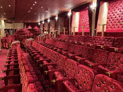 Eleganter Theatersaal auf der Carnival Venezia. Rote Samtsitze mit Gold-Details, Beistelltischen und Getränkehaltern. Roter Teppich und dekorative Säulen. Kein Publikum.