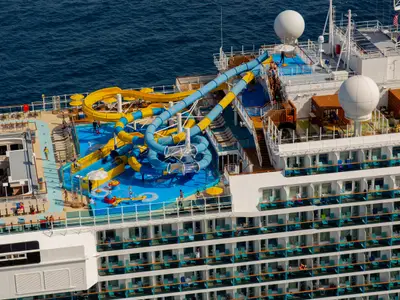 Vogelperspektive auf das Oberdeck der Carnival Venezia. Mehrere gelbe und blaue Wasserrutschen schlängeln sich über ein blaues Deck mit Pools und Kinderspielbereich. Zahlreiche Passagiere sind zu sehen, einige auf den Rutschen, andere auf Liegestühlen oder Balkonen.