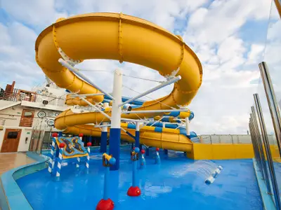 Eine weitläufige Wasserlandschaft auf dem Deck der Carnival Venezia mit mehreren gelben und blauen Wasserrutschen, einem Kinder-Sprühbereich mit bunten Spielgeräten und einem hellblauen Boden. Im Hintergrund ist die weiße Struktur des Schiffes und ein bewölkter Himmel zu sehen.