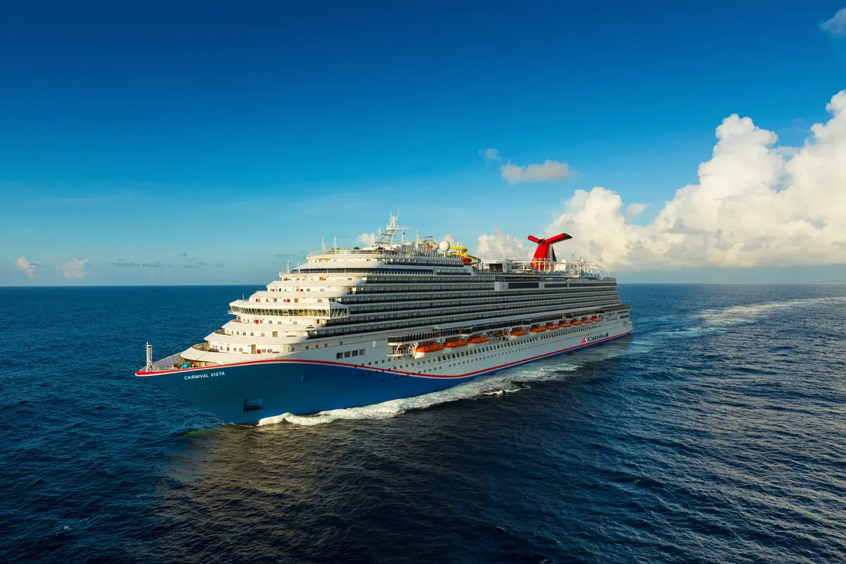 Eine Drohnenansicht der modernen Carnival Vista, die bei Tageslicht auf dem weiten blauen Meer fährt und eine weiße Spur hinter sich herzieht. Der Himmel ist teilweise bewölkt.
