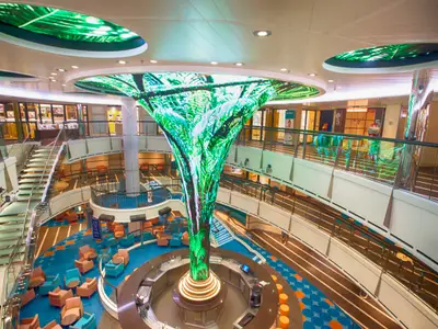 Weitwinkelaufnahme des mehrstöckigen Atriums der Carnival Vista. Eine große, baumartige LED-Skulptur mit Dschungelmotiven dominiert den Raum über einer runden Bar. Komfortable Sitzbereiche mit bunten Sesseln und eine geschwungene Treppe sind sichtbar. Die Decke zeigt ebenfalls grüne Lichtinstallationen. Das Atrium ist modern und hell beleuchtet.