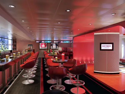 Moderne Sportbar auf der Carnival Vista in lebhaftem Rot. Eine lange Theke mit Barhockern und Hochtischen ist zu sehen. Zahlreiche Bildschirme zeigen Sport. Ein 'SKYBOX SPORTS BAR' Schild leuchtet.