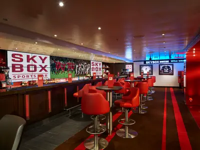 Eine lebhafte Sportsbar an Bord der Carnival Vista mit einer langen Bar, vielen roten Barhockern und Tischen. Mehrere große Bildschirme zeigen Sportereignisse und das Logo 'SKY BOX SPORTS BAR'. An den Wänden hängen gerahmte Trikots. Der Raum ist in Rot- und Brauntönen gehalten mit blauen Akzentlichtern.